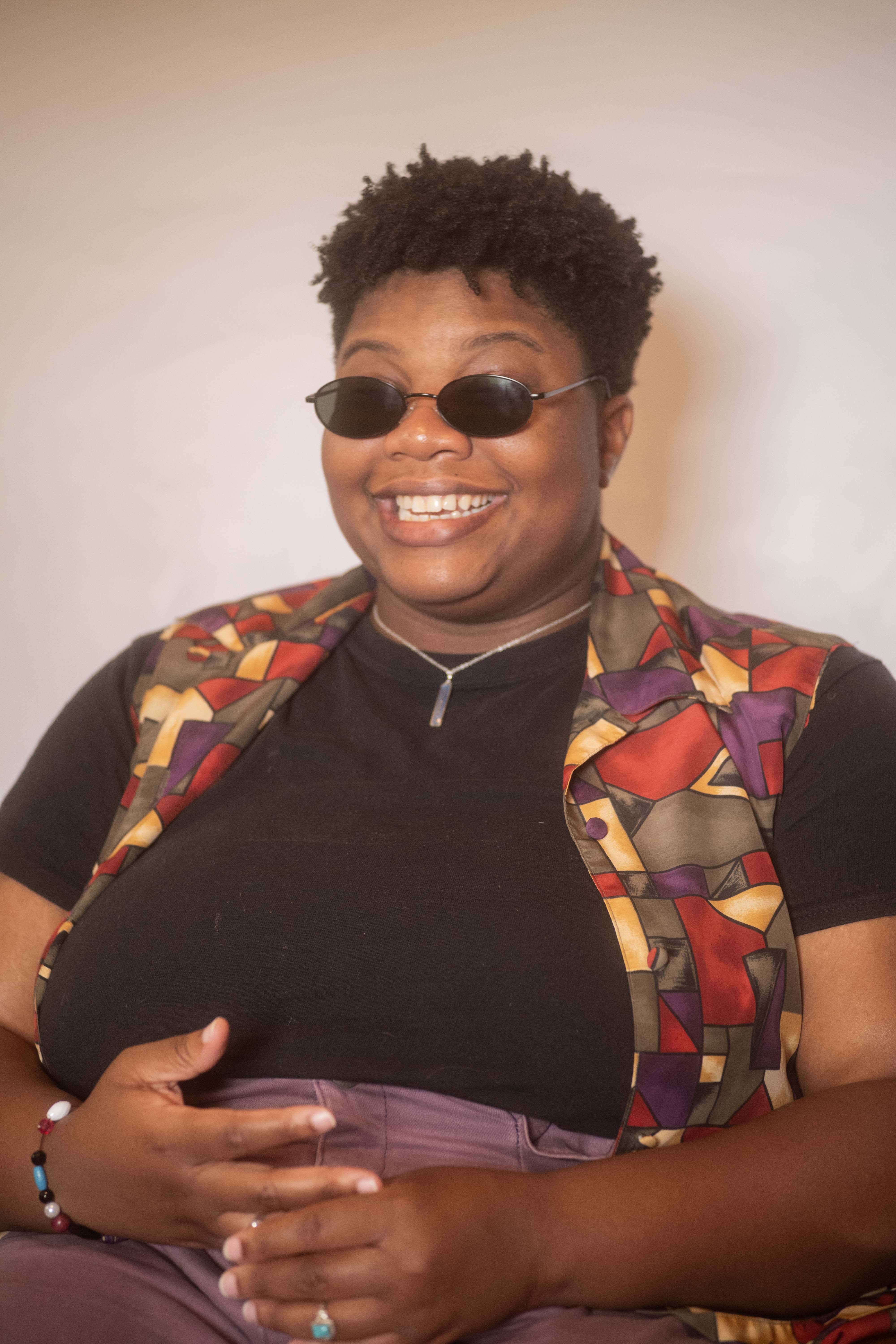 Kameron smiles cheekily in a colorful outfit and curly cropped haircut, with black oval sunglasses shielding their sensitive eyes.