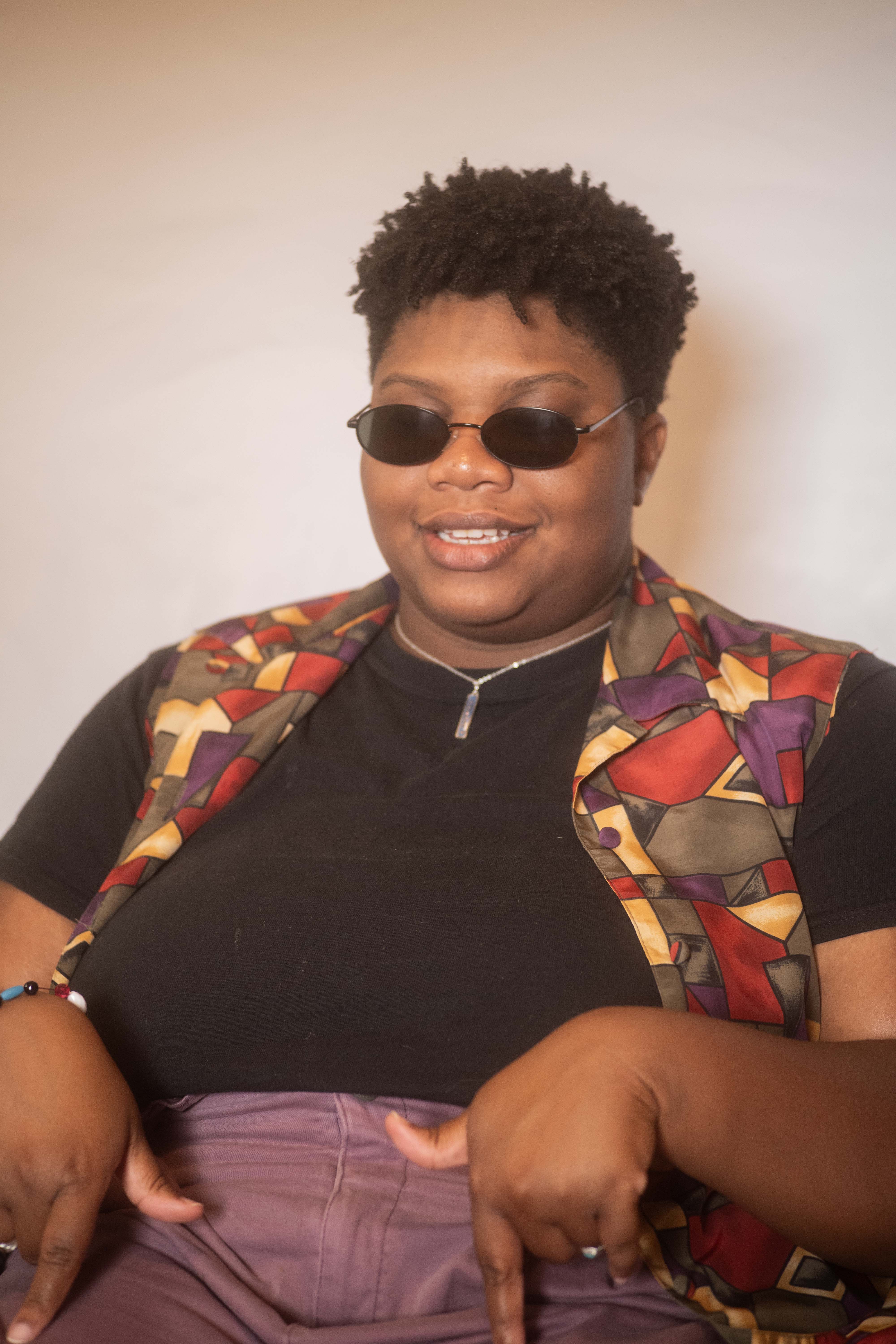 Kameron gestures at her lap with her thumb and forefinger in a colorful outfit and curly cropped haircut, with black oval sunglasses shielding her sensitive eyes.