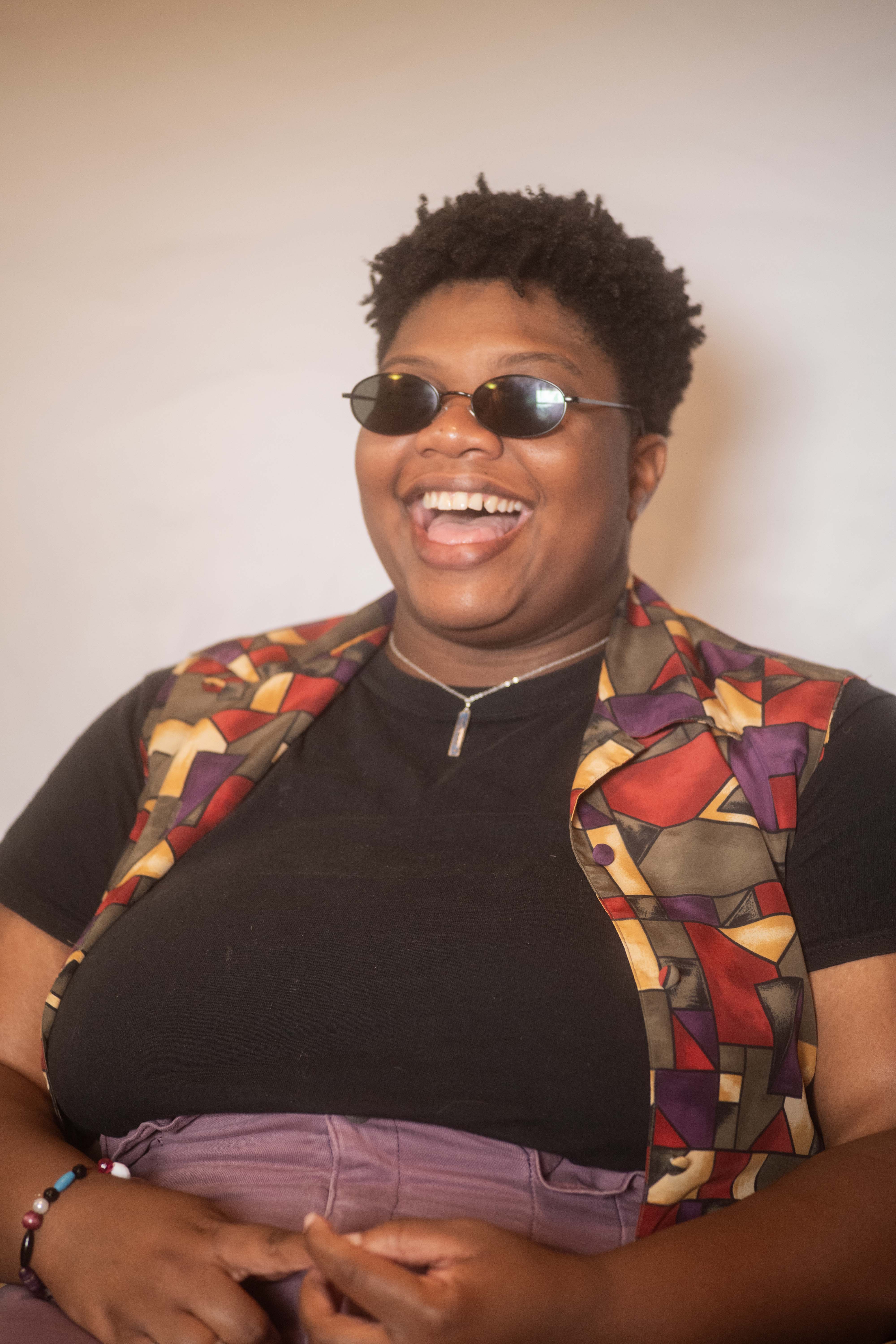 Kameron laughs boisterously in a colorful outfit and curly cropped haircut, with black oval sunglasses shielding his sensitive eyes.