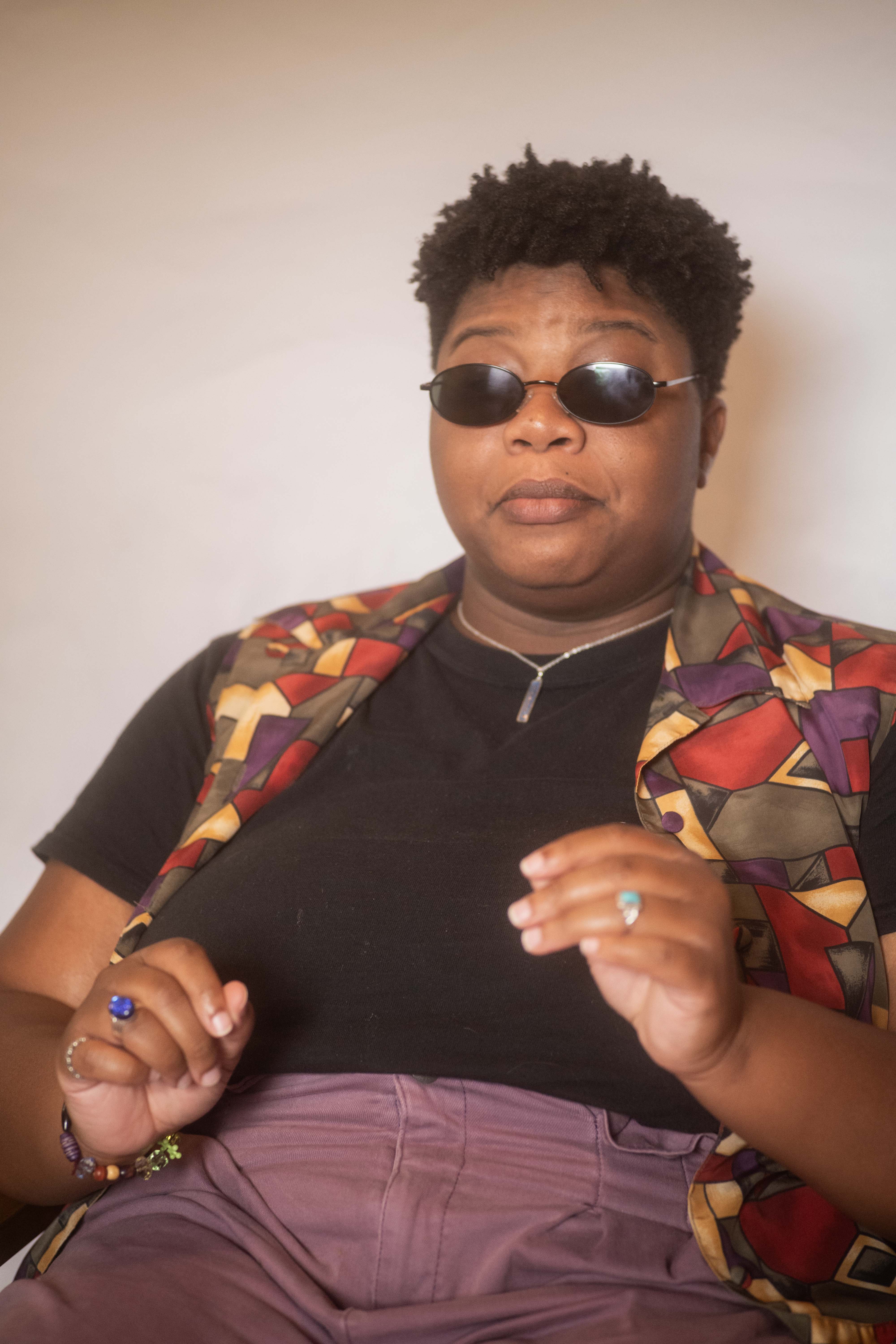 Kameron is caught mid-sentence in a colorful outfit and curly cropped haircut, with black oval sunglasses shielding their sensitive eyes.