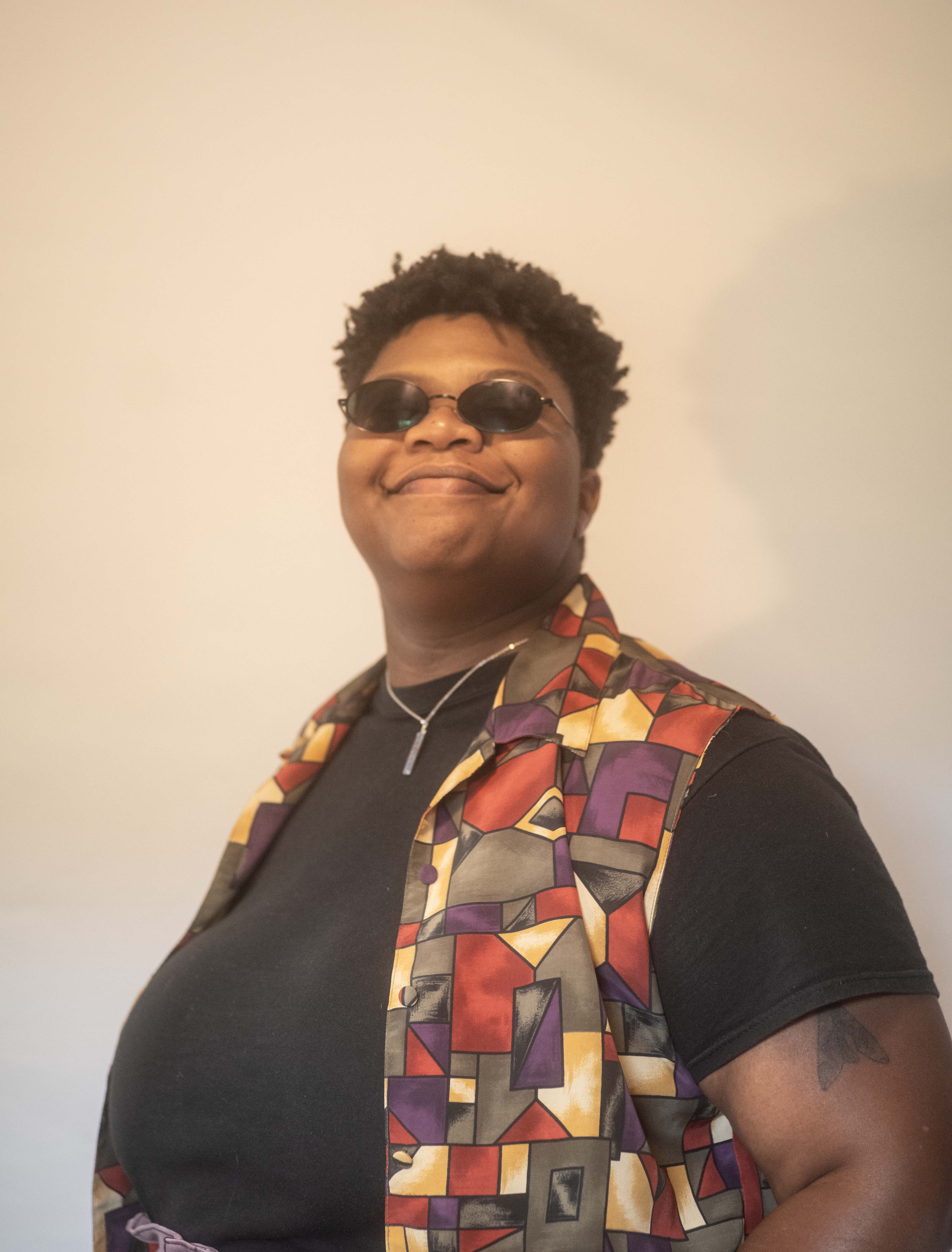 Kameron grins into the camera in a colorful outift and curly cropped haircut, with black oval sunglasses on.