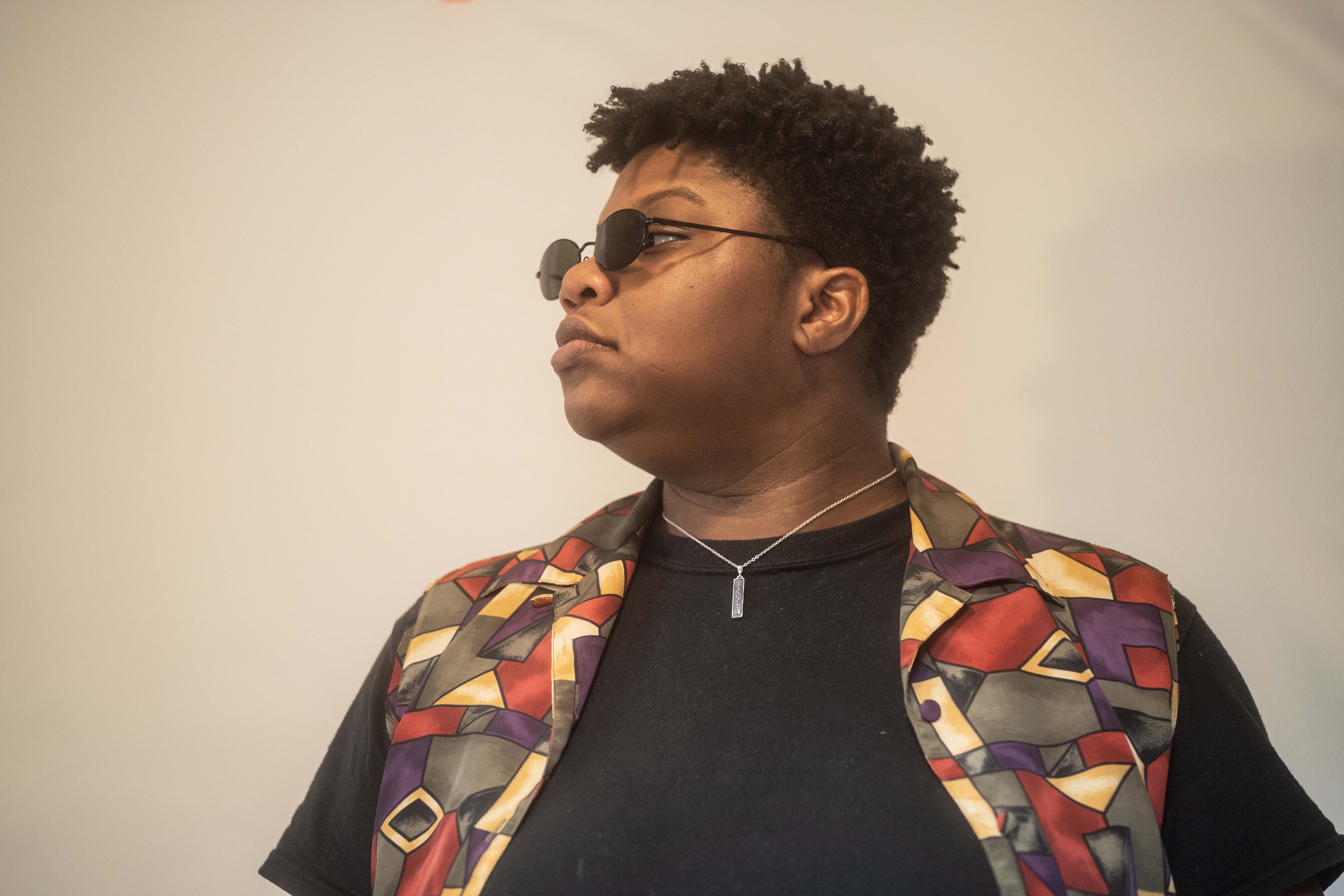 Kameron's left-side profile shot, looking serious in a colorful outfit and curly cropped haircut, black oval sunglasses on.